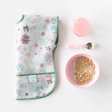 A folded floral bib, Bumkins Silicone Grip Bowl: Pink with cereal and milk, a pink cup, and pink spoon neatly placed on a white background.