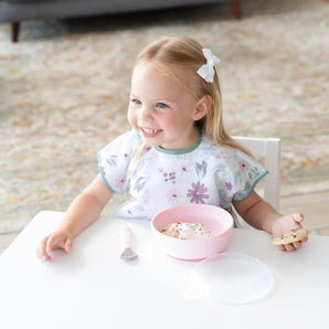 A happy toddler in a floral bib holds a cookie and enjoys ice cream from a Bumkins Silicone Grip Bowl: Pink at a table.