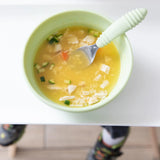 Bumkins Silicone Grip Bowl: Sage with chicken soup and a green spoon sits on a high chair tray; a child's feet are visible below.