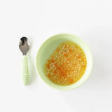 Bumkins Silicone Grip Bowl: Sage, filled with soup and pasta, sits on a white background next to a matching spoon.