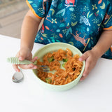 A child in a colorful bib eats pasta from a Bumkins Silicone Grip Bowl: Sage with a fork at a white table.