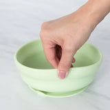 A hand holds a Bumkins Silicone Grip Bowl: Sage, demonstrating the suction cup bottom on a marble surface.