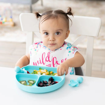 A toddler with pigtails enjoys a meal from a Bumkins Silicone Grip Dish: Blue, with sections of fruits and veggies, at a white table.