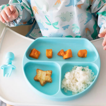 A baby in a dinosaur bib enjoys rice, sweet potato, and a star treat from a Bumkins Silicone Grip Dish: Blue with a blue fork nearby.