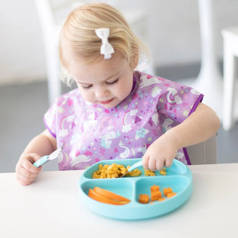 A toddler in a purple bib enjoys macaroni and veggies from a Bumkins Silicone Grip Dish: Blue with a fork at a white table.