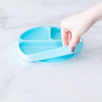 A hand holds a Bumkins Silicone Grip Dish: Blue, demonstrating the suction cup bottom on a marble surface.