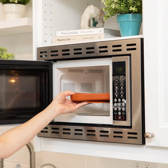 A person places a Bumkins Silicone Grip Dish: Clay inside an open microwave.