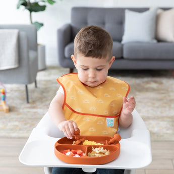 A toddler enjoys a meal from Bumkins Silicone Grip Dish: Clay in a high chair, with a cozy gray sofa in the background.