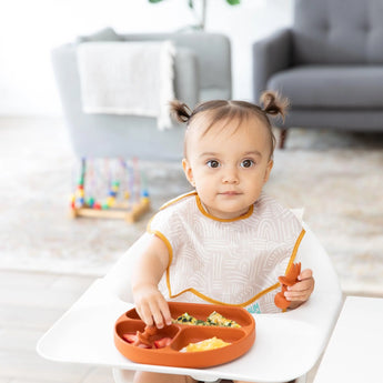 A toddler with pigtails in a high chair enjoys a meal from Bumkins Silicone Grip Dish: Clay, wearing a patterned bib in a cozy living room.