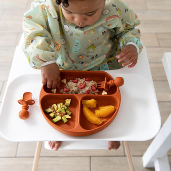 A toddler in a high chair eats oatmeal with berries, oranges, and avocado from Bumkins Silicone Grip Dish: Clay with their fingers.