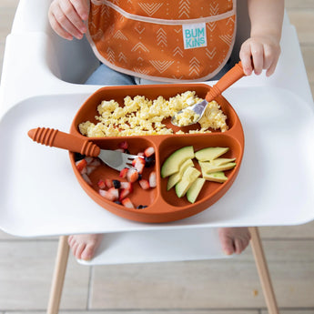 Close-up of a child in a high chair, wearing an orange bib, enjoying eggs, avocado, and fruit from Bumkins Silicone Grip Dish: Clay.
