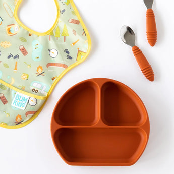 Bumkins Silicone Grip Dish: Clay sits next to a camping-themed bib and matching utensils on a white background.