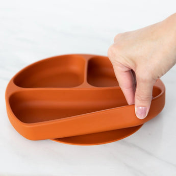 A hand holds a Bumkins Silicone Grip Dish: Clay, demonstrating the suction cup bottom on a marble surface.
