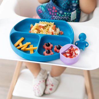 Close-up of a toddler in a high chair with a Bumkins Silicone Grip Dish: Dark Blue with pasta and cheese, and purple cup of fruit and yogurt.