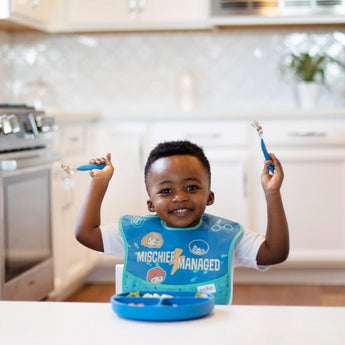 A happy toddler in a blue bib eagerly holds cutlery, ready to eat from a Bumkins Silicone Grip Dish: Dark Blue in a kitchen.