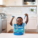 A smiling child wearing a blue bib holds Bumkins Spoon + Fork: Dark Blue above a matching plate on a white table in a bright kitchen.