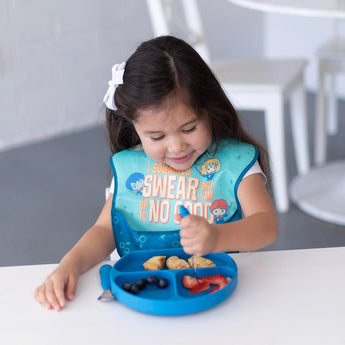 A smiling young girl in a blue bib enjoys fruit and snacks from a Bumkins Silicone Grip Dish: Dark Blue with utensils at a white table.