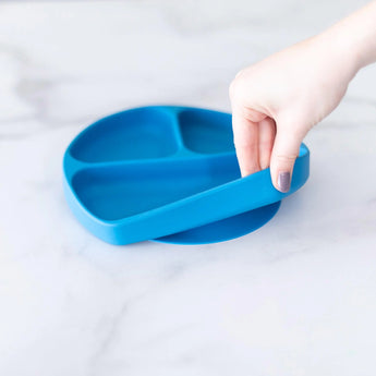 A hand holds a Bumkins Silicone Grip Dish: Dark Blue, demonstrating the suction cup bottom on a marble surface.