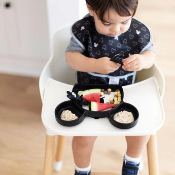 A toddler in a high chair wearing a black bib enjoys oatmeal, fruit, and cereal from a Bumkins Silicone Grip Dish: Mickey Mouse.