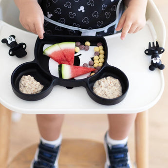 Close-up of a child in a high chair enjoying watermelon & cereal from a divided plate with Bumkins Silicone Chewtensils®: Mickey Mouse nearby.