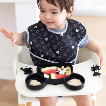 A toddler in a high chair uses Bumkins Silicone Chewtensils®: Mickey Mouse and a Mickey Mouse-shaped plate for baby-led weaning.