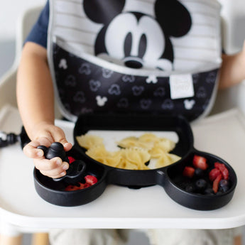 Close-up of a toddler in a high chair enjoying bowtie pasta and berries from a Bumkins Silicone Grip Dish: Mickey Mouse with a spoon.