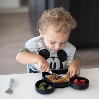 A toddler joyfully eats strawberries, grapes, and breaded food from a Bumkins Silicone Grip Dish: Mickey Mouse using a gray fork at a table.