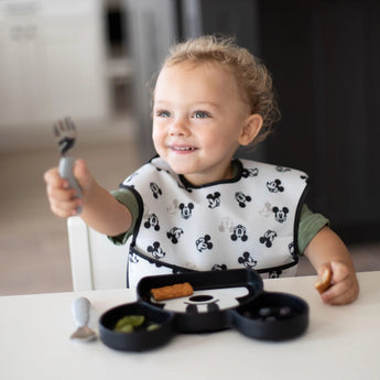A curly-haired toddler with a Bumkins Silicone Grip Dish: Mickey Mouse holds a fork at a white table, wearing a matching bib.