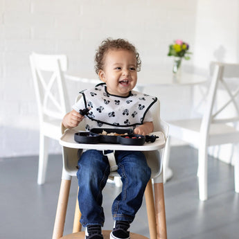 A toddler in a high chair, smiling with food in a Bumkins Silicone Grip Dish: Mickey Mouse, wearing jeans and a patterned bib.