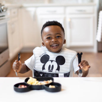 A toddler at a white table in a kitchen eagerly holds utensils over a Bumkins Silicone Grip Dish: Mickey Mouse with food.