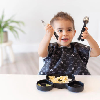 A smiling child eagerly grips a fork and spoon, ready to enjoy pasta from a Bumkins Silicone Grip Dish: Mickey Mouse at a white table.