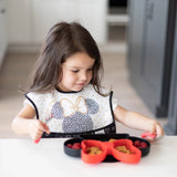 A young girl with long hair sits at a white table, enjoying food with a red fork from a Bumkins Silicone Grip Dish: Minnie Mouse.