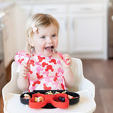 A toddler wearing a pink bib in a high chair eats from a Bumkins Silicone Grip Dish: Minnie Mouse with a red spoon and fork.