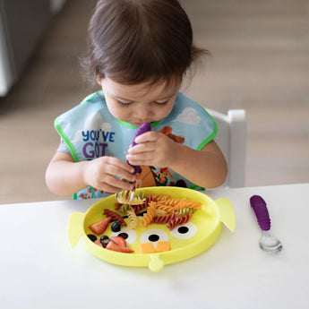 A toddler self-feeds from a Bumkins Silicone Grip Dish: Toy Story Alien with purple utensils at a white table.