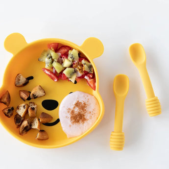 Bumkins Silicone Grip Dish: Winnie The Pooh holds fruit, toast, and yogurt near two yellow baby spoons on a white background.