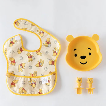 Bumkins Silicone Grip Dish: Winnie The Pooh sits near matching utensils and a yellow bear-print bib on a white background.