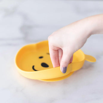 A hand holds a Bumkins Silicone Grip Dish: Winnie The Pooh, demonstrating the suction cup bottom on a marble surface.