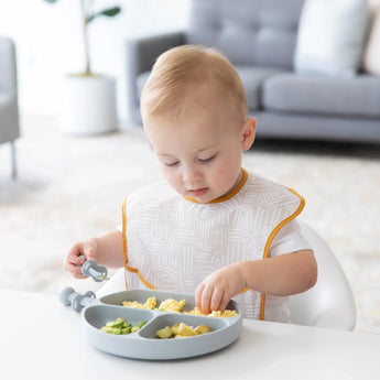 A toddler enjoys a meal from a Bumkins Silicone Grip Dish: Gray with gray toddler utensils at a white table in a bright living room.