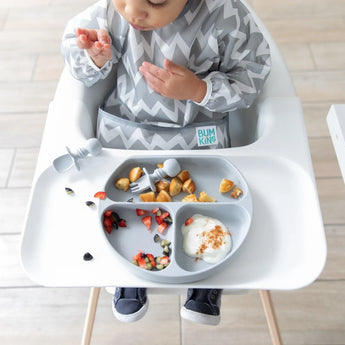 A baby in a high chair wearing a gray zig-zag bib enjoys food from a Bumkins Silicone Grip Dish: Gray with matching utensils.