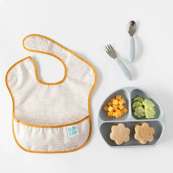 Bumkins Silicone Grip Dish: Gray cheese cubes, cucumber slices, and sandwiches near gray utensils and a beige bib on a white background.