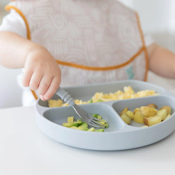 Close-up of a child in a beige bib enjoying veggies with a fork from a Bumkins Silicone Grip Dish: Gray at a white table.