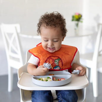 A curly-haired toddler in an orange bib smiles in a high chair, enjoying food from a Bumkins Silicone Grip Dish: Gray with a toddler fork.