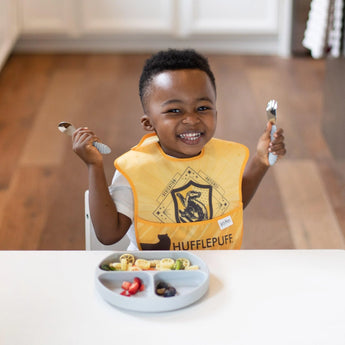 A child in a yellow bib beams while holding utensils at a white table near a Bumkins Silicone Grip Dish: Gray with pasta and fruit.