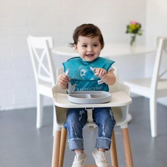 A toddler in a high chair wearing a blue bib joyfully holds a spoon near a Bumkins Silicone Grip Dish: Gray in a bright room.