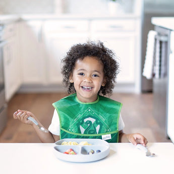 A curly-haired child in a green bib beams at a white table, enjoying a meal from a Bumkins Silicone Grip Dish: Gray in a bright kitchen.