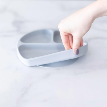 A hand holds a Bumkins Silicone Grip Dish: Gray, demonstrating the suction cup bottom on a marble surface.
