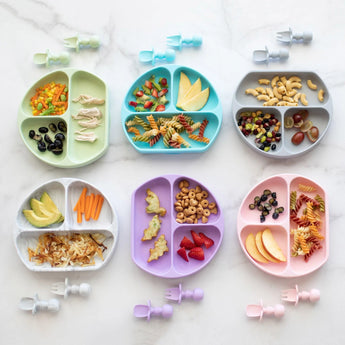 Six toddler plates, including a Bumkins Silicone Grip Dish: Gray, with snacks and matching utensils are neatly arranged on a marble surface.