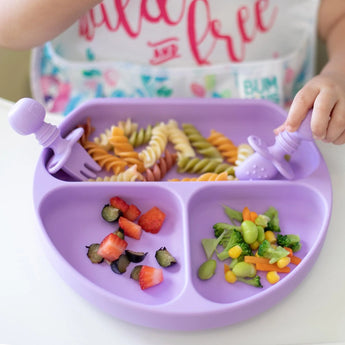 Close-up of a child enjoying food from a Bumkins Silicone Grip Dish: Lavender with matching utensils while wearing a colorful bib.