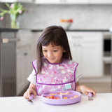 A young girl in a colorful bib sits at a white table with a purple plate, holding Bumkins Spoon + Fork: Lavender.