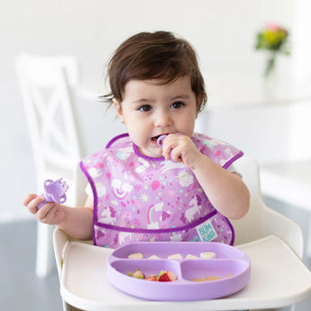 A baby wearing a purple bib in a high chair enjoys a meal from a Bumkins Silicone Grip Dish: Lavender with matching utensils.